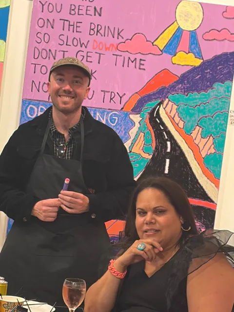 Two people smiling in front of a large painted work in the gallery