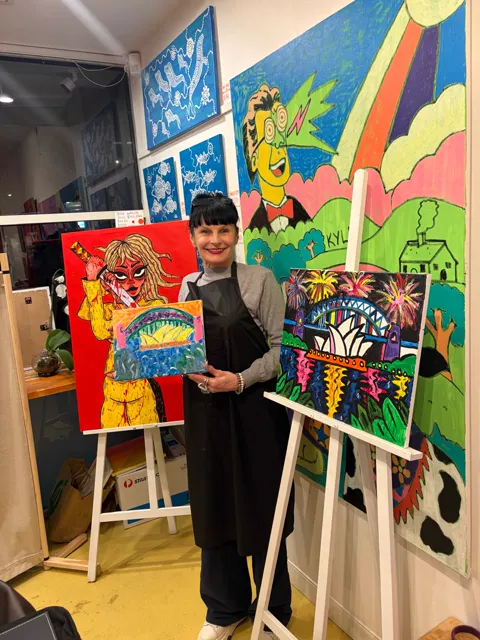 A guest holding up her finished painting next to the large fireworks painting